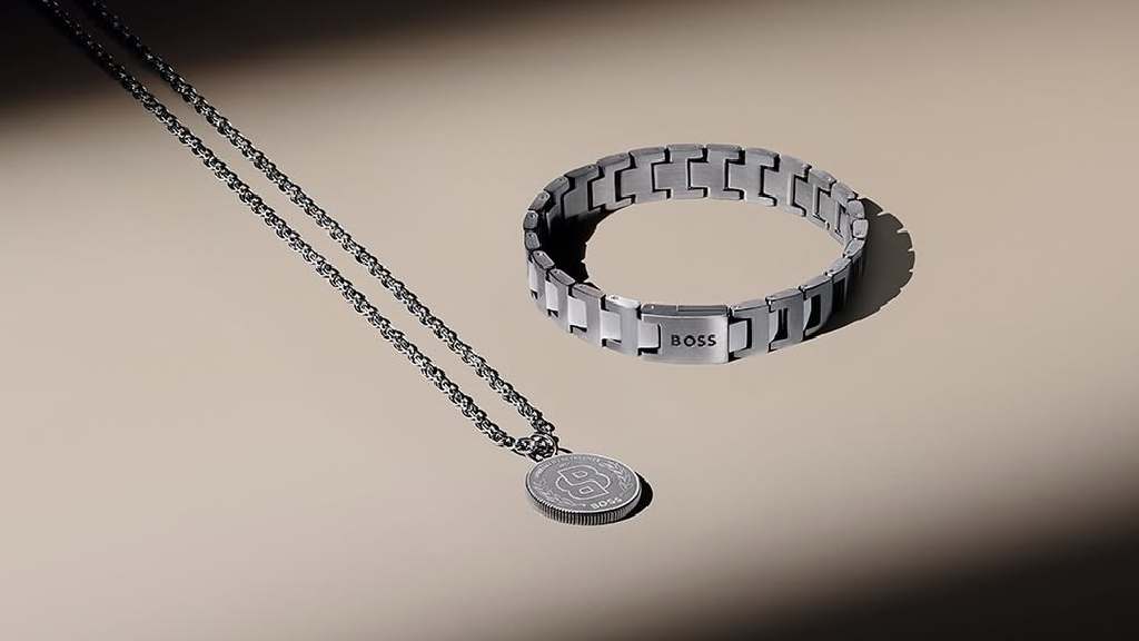 Foto de colar masculino ao lado de pulseira de aço prata ambos da coleção Boss Jewelry.